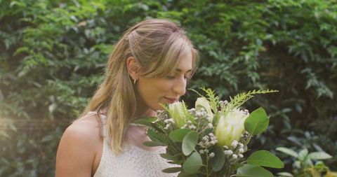 Woman smelling white bouquet in sunlit garden wearing sleeveless lace dress and gold hoops