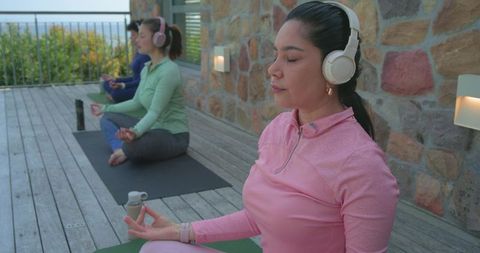 Women meditating with headphones on coastal deck practicing guided yoga and mindfulness
