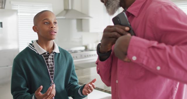 Father is holding a smartphone while son is gesturing during a serious conversation in a modern kitchen. Ideal for use in articles about family dynamics, parenting challenges, and communication within families.