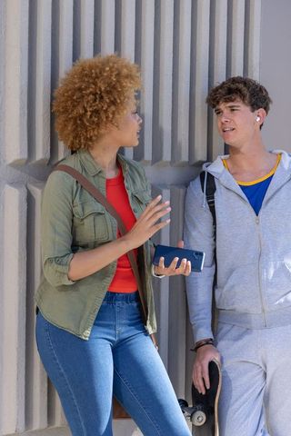 Diverse teens chatting and leaning on urban grooved concrete wall with skateboard and smartphone