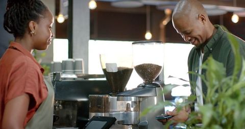 African american barista serving smiling customer using mobile pay, modern coffee counter