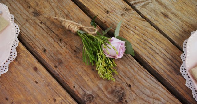 A single pink rose with greenery tied with twine lies on a rustic wooden table, with copy space. The setting suggests a simple, natural decoration, for a wedding or a romantic event.
