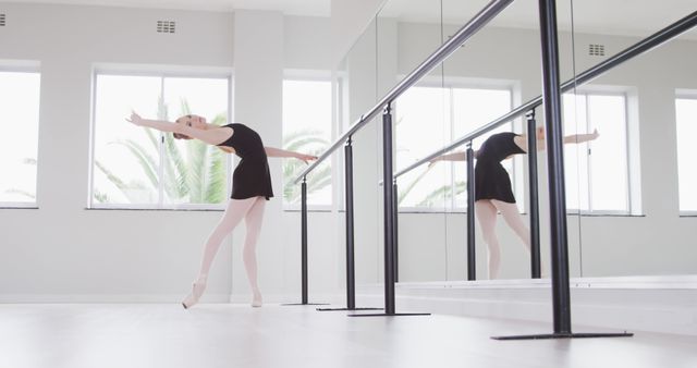 Ballet dancer practicing gracefully at a barre in a bright, spacious studio with large windows and natural light. The studio features mirrors, enhancing the dancer's concentration on form and technique. Light wooden floor and neutral decor create a serene atmosphere, perfect for dance practice. This image is ideal for promoting ballet classes, dance schools, fitness ads, and any material related to performing arts and physical wellness.