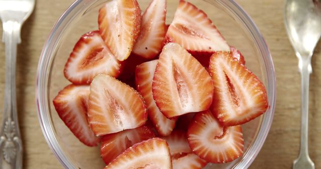 Close-up of fresh strawberry halves in a clear bowl on a wooden table. Ideal for use in healthy eating campaigns, food blogs, recipes, and grocery store advertisements emphasizing fresh, organic produce.