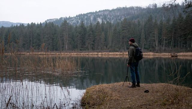 Solitary hiker surveying tranquil forest lake shore with tripod and backpack