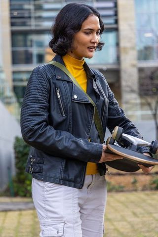 Carrying skateboard in urban courtyard wearing leather jacket and yellow turtleneck