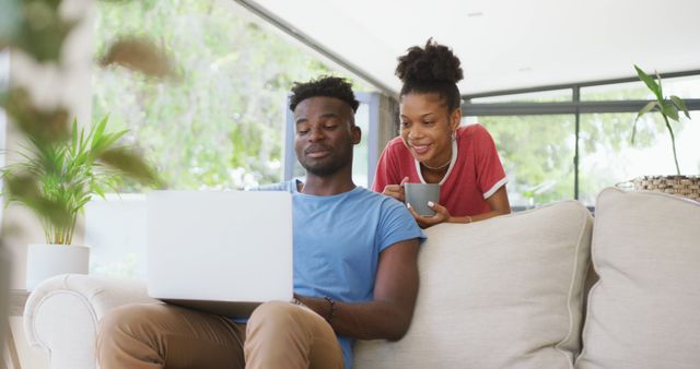 Image of happy african american couple on sofa with coffee and laptop. love, relationship, togetherness and spending time with technology at home.