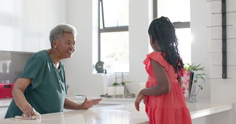 Grandmother smiling while wiping counter and granddaughter talking in sunlit modern kitchen