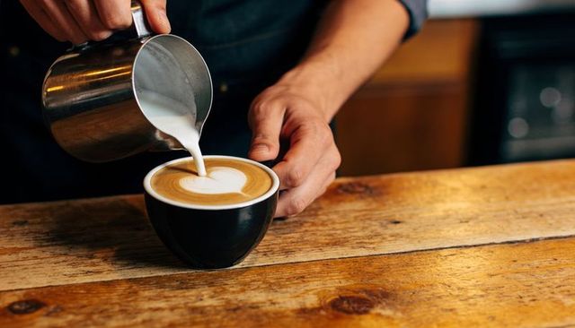 Barista pouring heart latte art over black cup on rustic wooden countertop