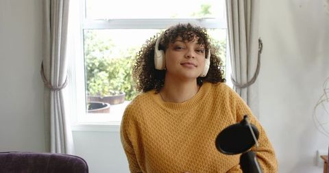 Young woman recording podcast at home by sunlit window wearing mustard sweater and headphones