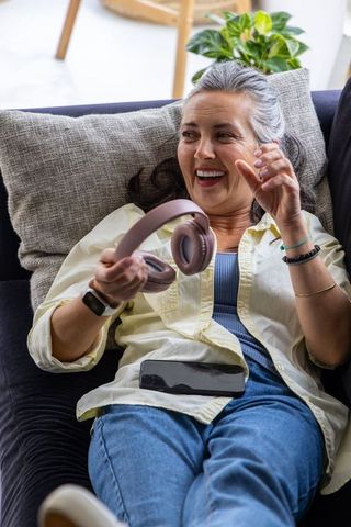 Gray-haired woman laughing while reclining on sofa holding headphones and smartphone