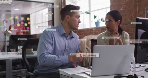 Colleagues collaborating at open-plan office desk with laptop, documents, data overlays