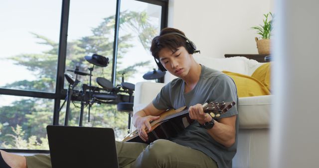 Asian boy wearing headphones playing guitar looking at the laptop at home. teenager lifestyle and living concept