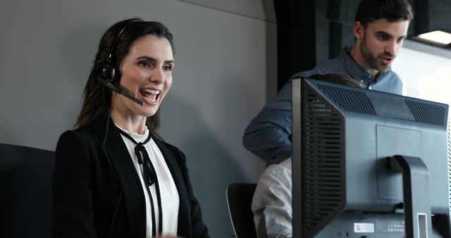A Caucasian woman wearing a headset is smiling as she works at her computer, with a young Caucasian man in the background focusing on his screen, both professionals in a business or call center environment. Their engagement and attire suggest a dynamic and customer-oriented workplace.
