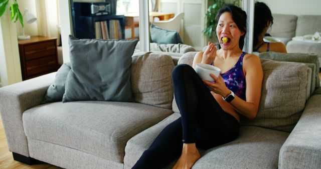 Woman lounging on couch while enjoying a healthy snack wearing fitness attire. Suitable for use in publications about healthy living, relaxation at home, fitness lifestyle, and casual comfort. Perfect for promoting lifestyle blogs, health food products, or home decor ideas.