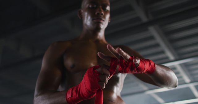 A strong male boxer is wrapping his hands with red bandages in a dark gym. He is focusing on preparing for intense training, highlighting his muscles and determination. This image can be used for fitness blogs, sports articles, motivational content, or advertisements for gyms and boxing gear.