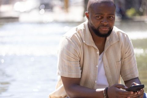 African american man sitting by sunlit waterfront holding smartphone in beige shirt
