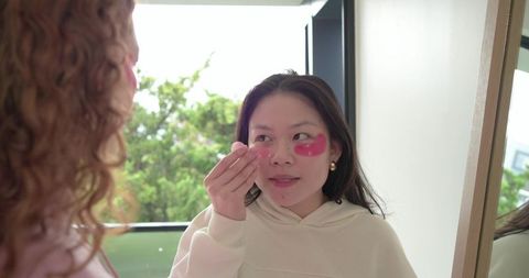 Friends applying pink hydrogel undereye patches by mirror near balcony for skincare routine