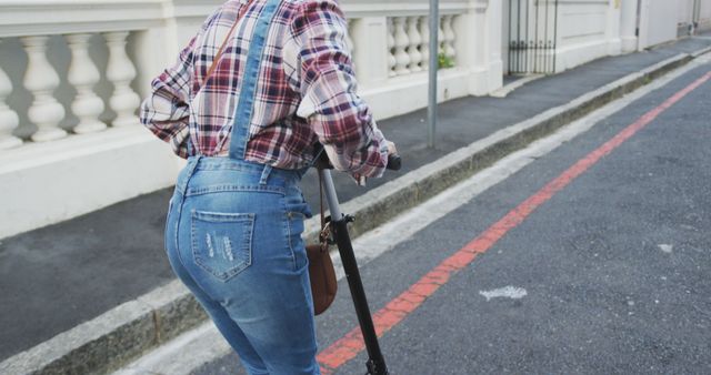 This image features a woman riding an electric scooter down an urban street. She is wearing a flannel shirt and denim overalls, suggesting a casual and trendy style. The street setting indicates city life and modern transportation. Ideal for marketing campaigns related to urban mobility, eco-friendly transportation, casual fashion, or city life experiences.