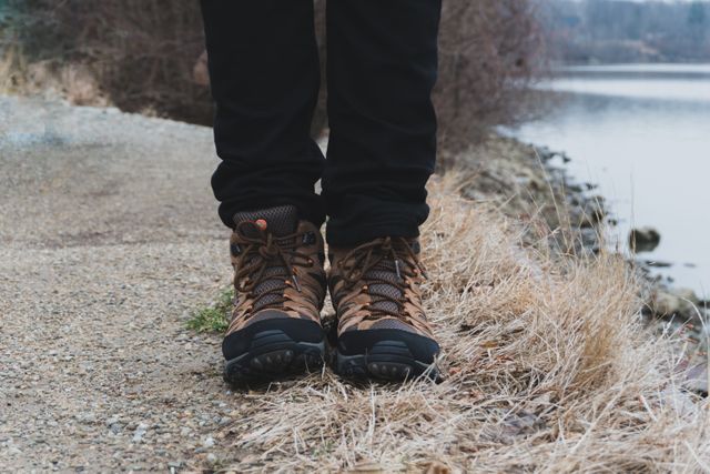 Picture of a hiker wearing brown boots standing by a lake on a gravel path. Surrounding dry grass and an overcast sky emphasize calm and isolation, making it ideal for mood pieces focusing on outdoor adventure, solitude, travel blogs, or promotional material for outdoor apparel brands.