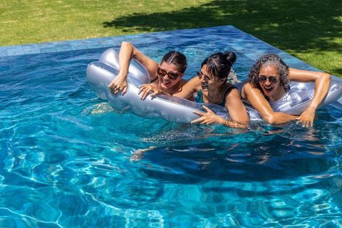 Three women laughing and leaning on silver inflatable pool float in sunny backyard