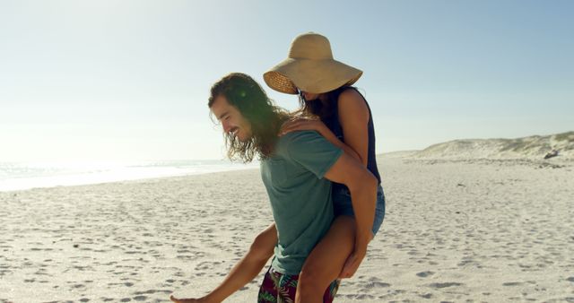 Young couple enjoying a playful piggyback ride on the sunlit sandy beach with clear skies and ocean in the background. Suitable for travel advertisements, summer holiday promotions, lifestyle blogs, and social media content related to outdoor activities and relationship goals.