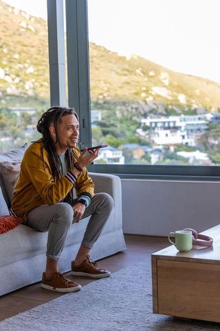Smiling african american man speaking into smartphone while sitting on gray sofa with hillside view