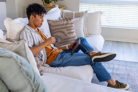 Young asian man reclining on sofa swiping credit card on tablet for online shopping