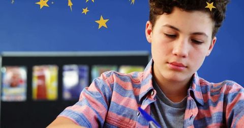 Teen Boy Writing at School Desk Concentrating on Homework with Blue Classroom Background