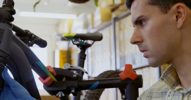 Man repairing bicycle with a focused expression, surrounded by tools and equipment in a well-lit workshop. Useful for illustrating concepts related to mechanical work, bicycle maintenance, and technical skills. It can also be used in promotional materials for cycling shops, repair services, or technical training programs.