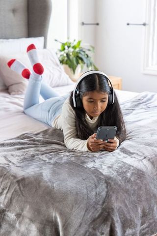 Indian girl lying on bed wearing headphones and using smartphone in bright bedroom