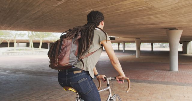 Depicts an urban cyclist commuting on a bicycle under a concrete bridge. Perfect for use in advertising urban lifestyles, transportation solutions, eco-friendly travel, active lifestyles, or youth culture. Ideal for blog posts or articles on cycling, urban commuting, or sustainable travel methods.