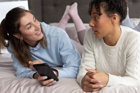 Multiracial women chatting on bed with smartphone, cozy relaxed friendship moment