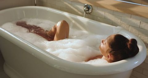 Woman Relaxing in Foamy Bathtub Sunlit by Window Blinds