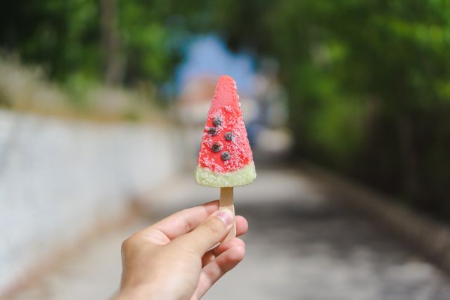 Close-up view of a hand holding a watermelon-shaped popsicle on a sunny day. Suitable for use in summer-themed content, advertisements for cold treats, and outdoor activity promotions.