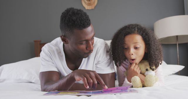 Father engages his daughter in a colorful storybook while lying on the bed together. The father points to illustrations, sparking excitement in his daughter who is holding a stuffed animal. Ideal for themes related to family bonding, fatherhood, parenting, and bedtime stories. Can be used in educational materials, advertisements for parenting products, or articles about literacy and family routines.
