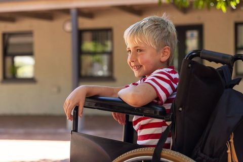 Smiling young boy in wheelchair leaning on armrest outdoors in sunlit courtyard wearing red shirt