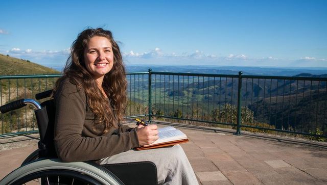 Smiling woman in wheelchair writing journal on mountain viewpoint with valley vista