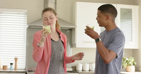 Diverse couple tasting green smoothies in bright kitchen wearing smartwatches