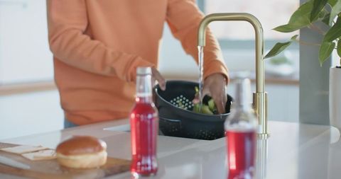 Man washing greens in colander under brass faucet on modern kitchen island with red drinks