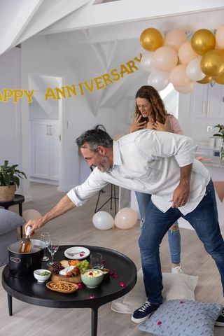 Middle-aged couple celebrating anniversary at home with wine, cheese board, pastel balloons