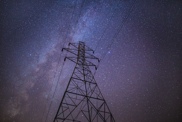 High-voltage power line tower stands tall against a mesmerizing starry night sky featuring the Milky Way. Great for illustrating themes related to energy, astronomy, engineering, and the blend of technology with nature. Ideal for websites, educational materials, articles on energy infrastructure, or cosmic-themed designs.