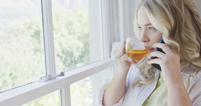 The image showcases a young woman enjoying a cup of tea while conversing on the phone next to a sunny window. With sunlight bathing the scene, this picture conveys a sense of calm and relaxation, making it ideal for themes related to morning routines, everyday lifestyle, or personal communication. Use this image for articles about remote work, relaxation tips, or personal well-being.
