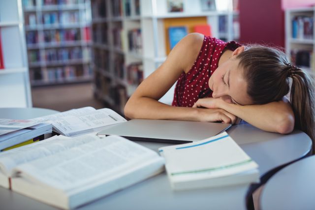 Young girl sleeping on desk in school library surrounded by open books and notebooks. Ideal for illustrating academic stress, student life, education challenges, and the importance of rest. Useful for articles, blogs, and educational materials focusing on student well-being, study habits, and mental health.