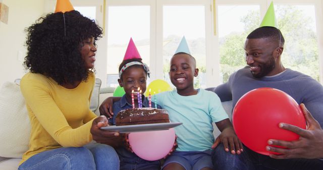 Happy family celebrating child's birthday at home. Mother holding a cake with colorful candles, while father and children are smiling and holding balloons. This can be used for advertising family products, birthday-themed materials, well-being, and party planning resources.