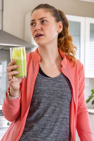 Young woman wearing coral jacket holding green smoothie and speaking in modern kitchen