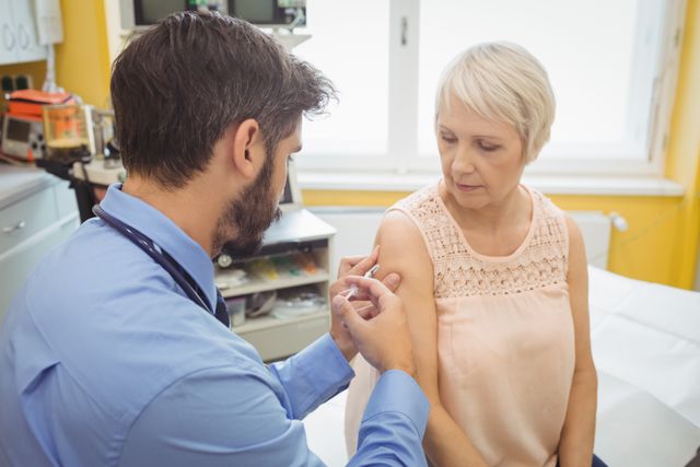 Male doctor giving an injection to a senior female patient at a hospital. The patient appears calm and is wearing a sleeveless top for easy access to her arm. This image is useful for illustrating healthcare services, vaccination programs, patient care, and medical treatments in hospitals and clinics.