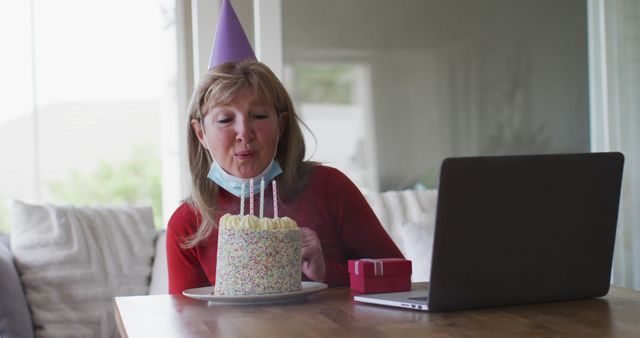 Senior woman wearing party hat blowing candles on a birthday cake while attending a virtual party on her laptop. Suitable for illustrating remote celebrations, pandemic social interactions, virtual gatherings, elderly technology use, and safety measures during health crises.