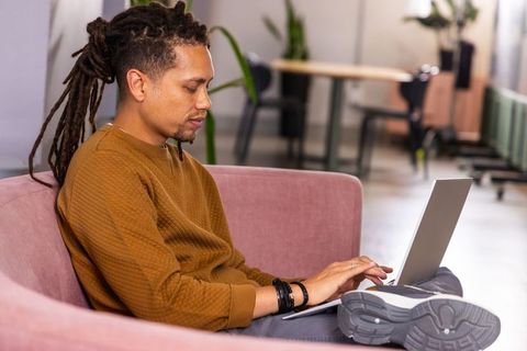 Young dreadlocked man using laptop on pink couch in modern coworking space