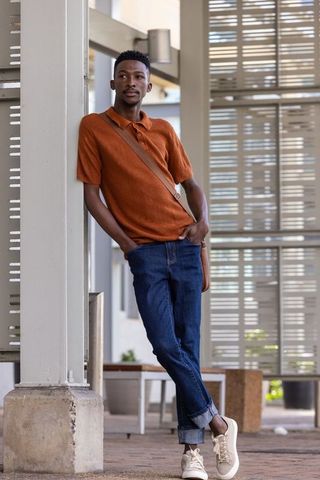 Young african man leaning on column wearing rust polo and crossbody bag in urban walkway